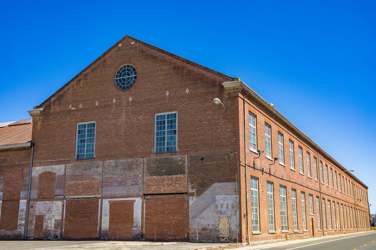 Exterior view of a decaying abandoned brick warehouse with tall windows and a clear blue sky.